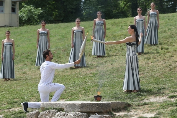 扮演最高女祭司的演員瑪麗·米娜（前右）點燃第一棒火炬手——希臘運動員、東京奧運會男子賽艇項目金牌得主斯特凡諾斯·恩圖斯科斯手中的火炬。新華社記者 趙丁喆 攝